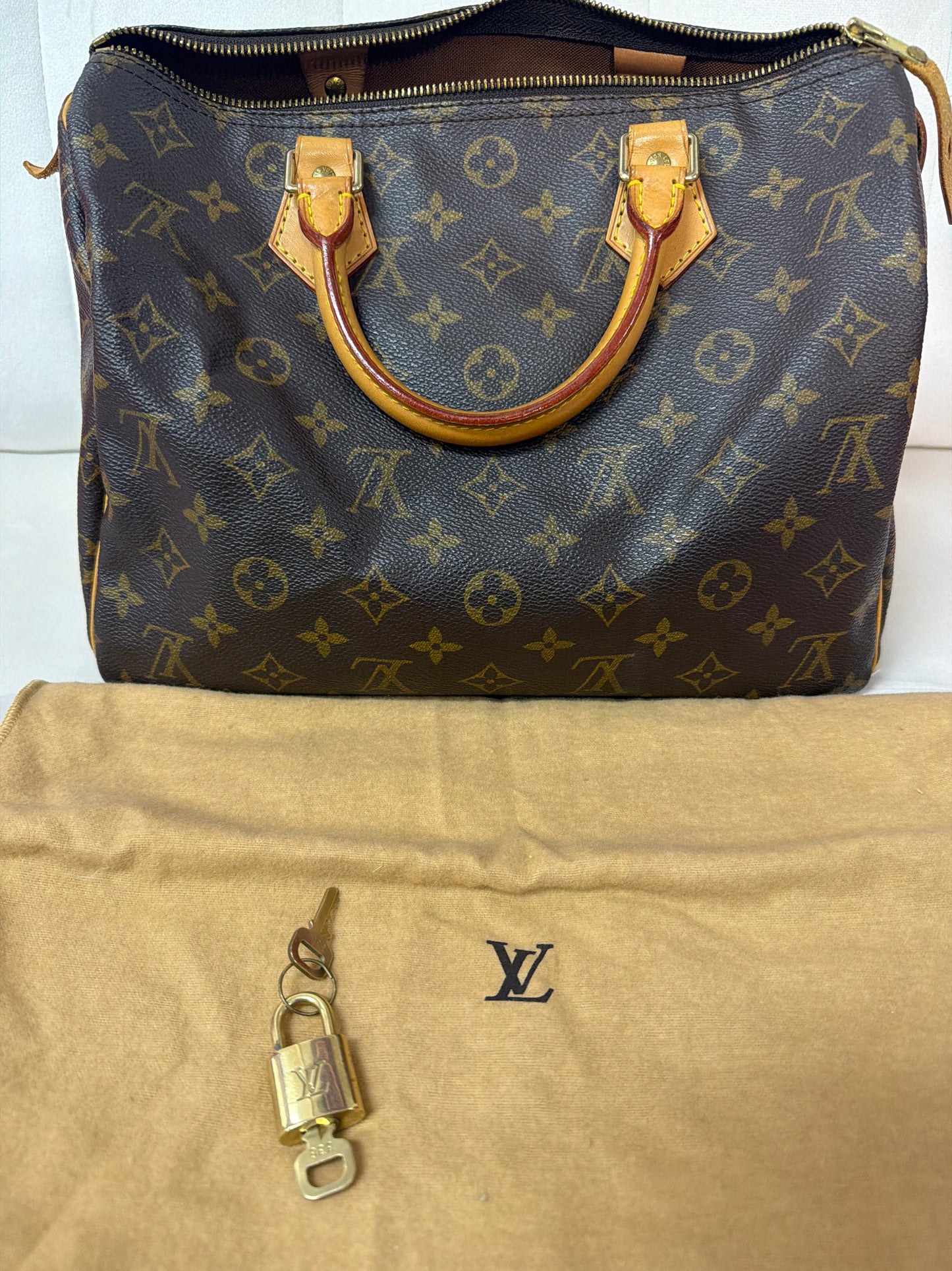 A Louis Vuitton monogram Speedy handbag in dark brown coated canvas with tan leather handles, shown above a tan dust bag with a small gold padlock and key.