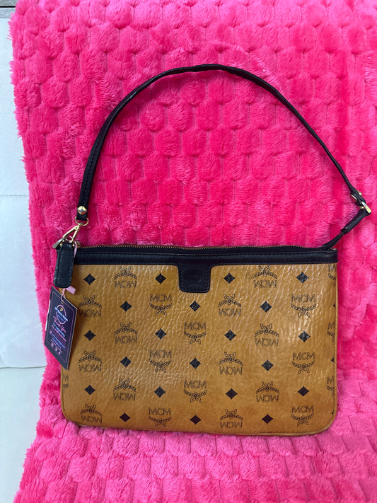 MCM monogram canvas shoulder bag in tan and black, featuring a top zipper, black shoulder strap, and a hanging brand tag, displayed on a textured hot pink background.