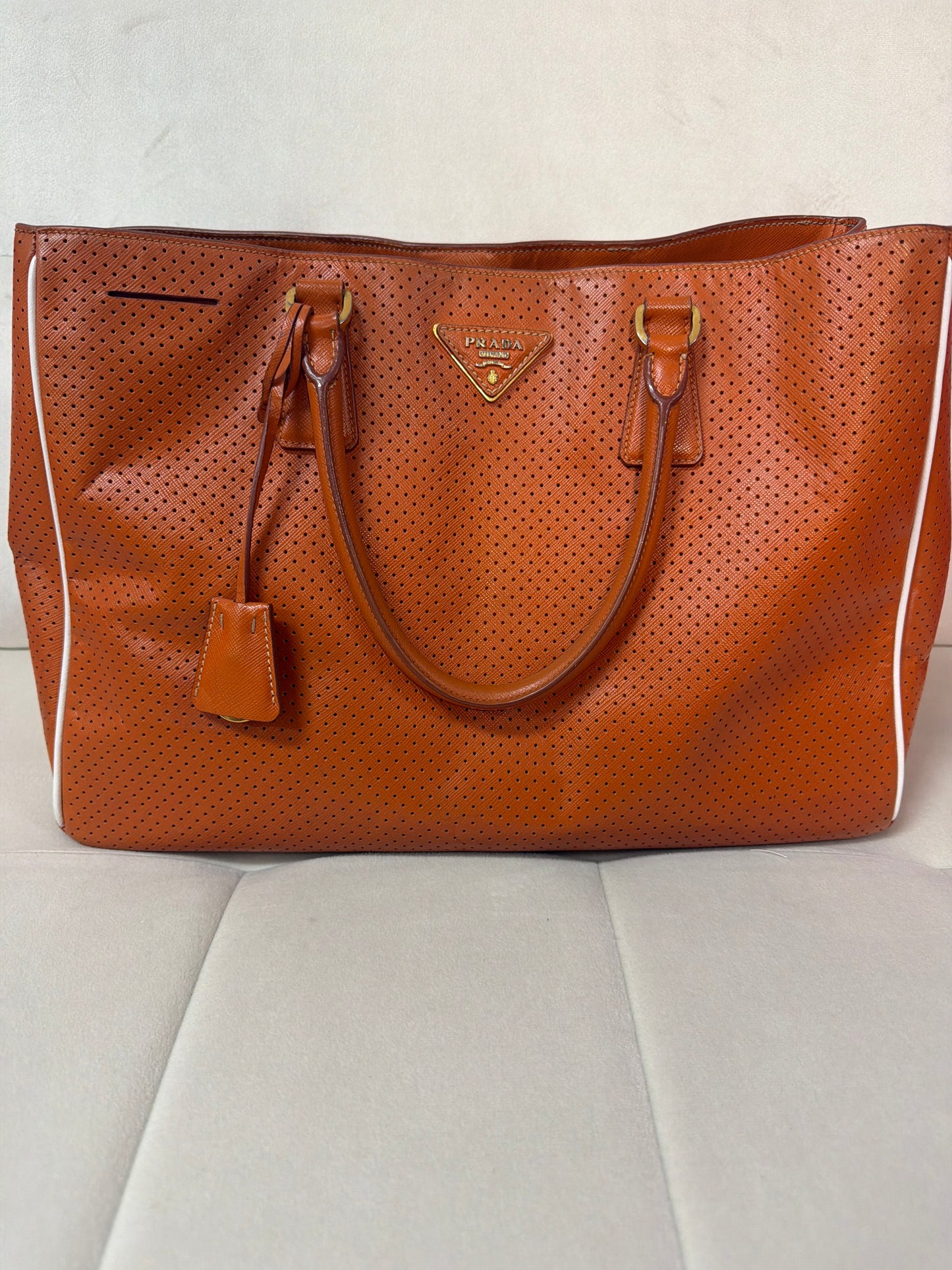 A Prada perforated leather tote bag in a warm orange , featuring dual top handles, a hanging leather tag, and the signature gold-tone triangular Prada logo plaque on the front, set against a light gray upholstered surface.