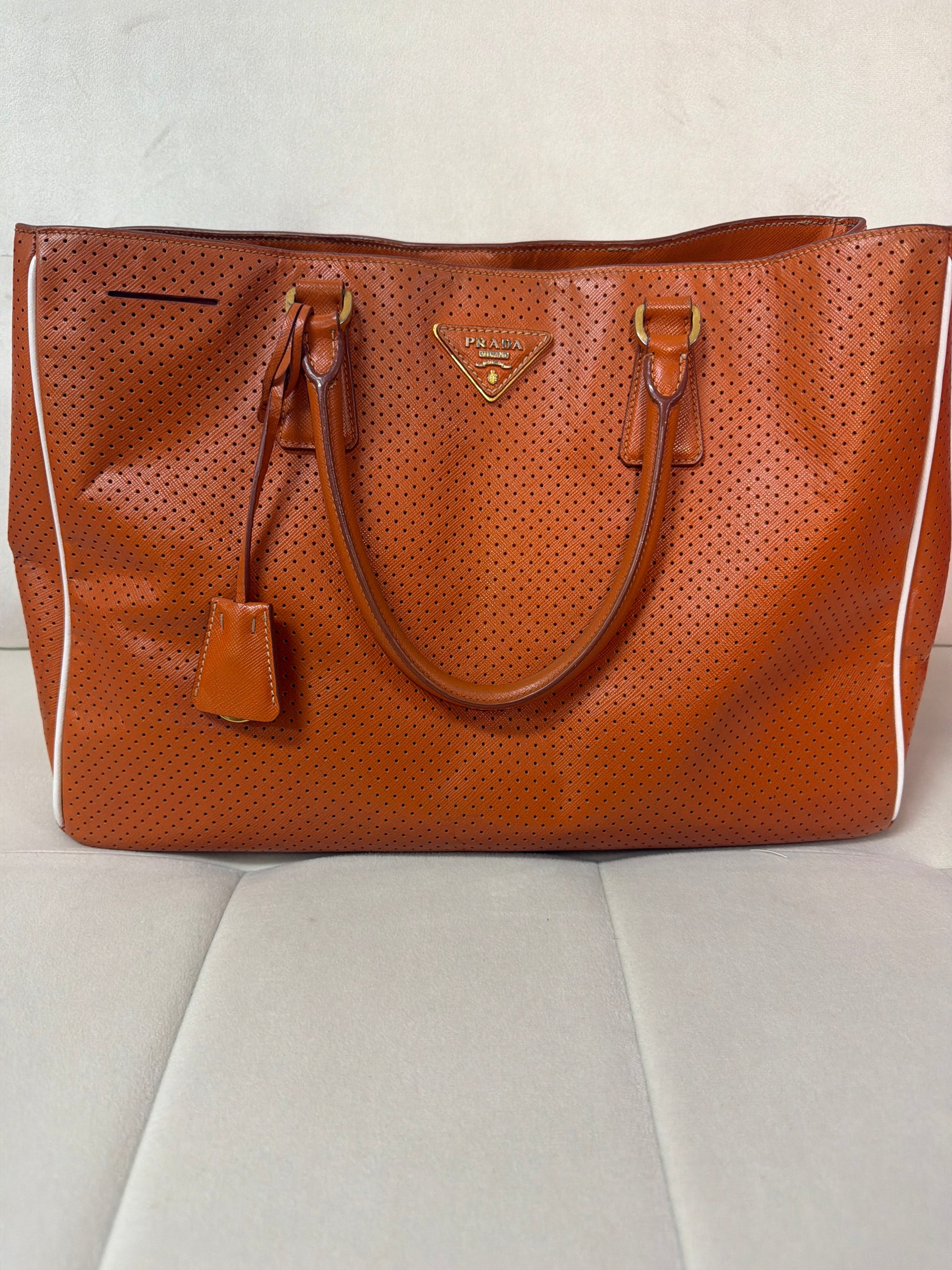 A Prada perforated leather tote bag in a warm orange , featuring dual top handles, a hanging leather tag, and the signature gold-tone triangular Prada logo plaque on the front, set against a light gray upholstered surface.